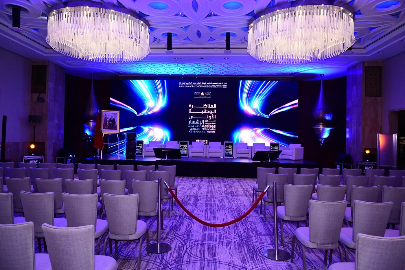 A conference hall with chandeliers, a blue LED stage and rows of white chairs.