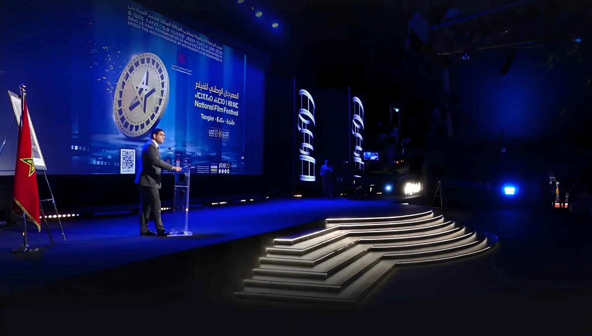 A speaker at a lectern on a blue-lit stage at the National Film Festival in Tangier, with lit steps in the foreground.