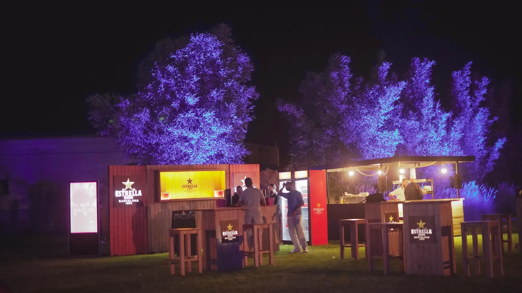 The bar zone at night: branded counters glowing under trees lit in violet.