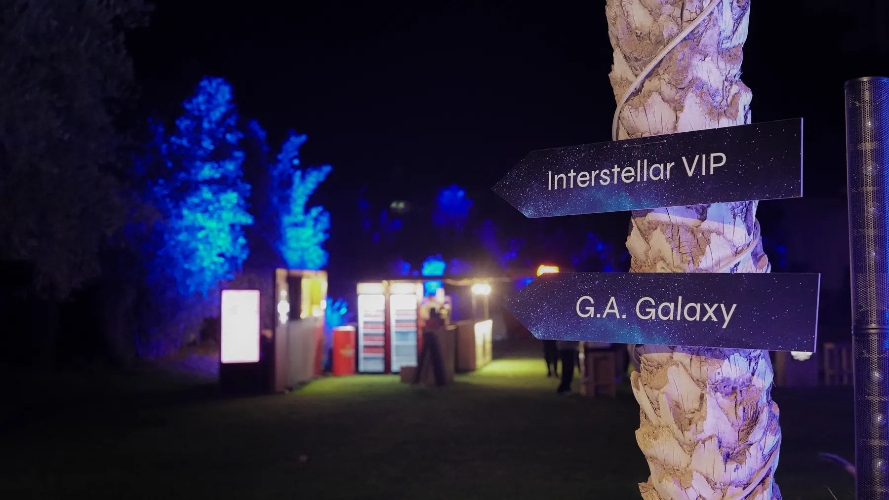 Wayfinding signpost on a palm trunk pointing to Interstellar VIP and G.A. Galaxy, with lit stands behind.